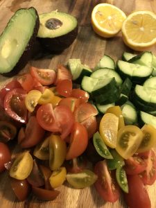 Tomatoes and cucumbers on a cutting board with avocado and lemons.