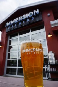 A glass of beer with Immersion Brewing in the background.