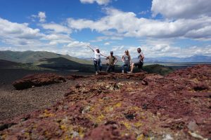 Four adults posing at Craters of the Moon National Monument with mountains and clouds in the background.