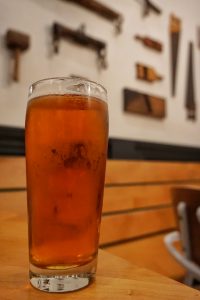 A glass of beer on a table with farmhouse tools on the wall in the background.