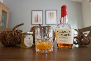 Crystal glass with a pumpkin spice hot toddy surrounded by decorative pumpkins, a bottle of whiskey, and a jar of honey.